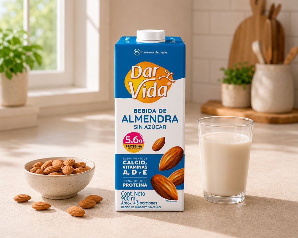 Empaque de Bebida de Almendra Sin Azúcar Dar Vida sobre una mesa clara, acompañado de un vaso con bebida de almendra y un bowl con almendras. Al fondo se observa una cocina iluminada con luz natural, plantas, utensilios y elementos de madera.